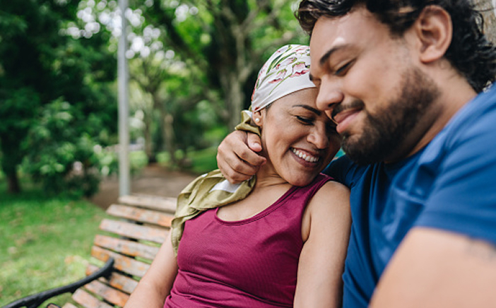 Smiling couple embraces on a park bench, sharing a moment of warmth during cancer care.