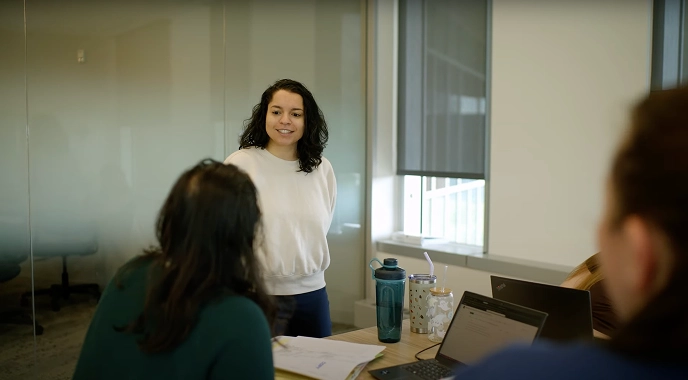 A woman stands in a meeting room, engaging with colleagues, sharing insights on cancer care stories.