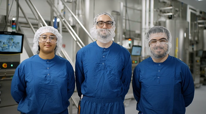 Three lab technicians in blue uniforms and hair nets stand together in a lab, contributing to cancer care advancements.