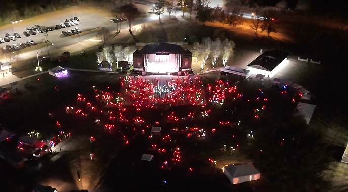 Aerial view of a nighttime event with a stage and crowd holding red lights, possibly related to a cancer care awareness event.